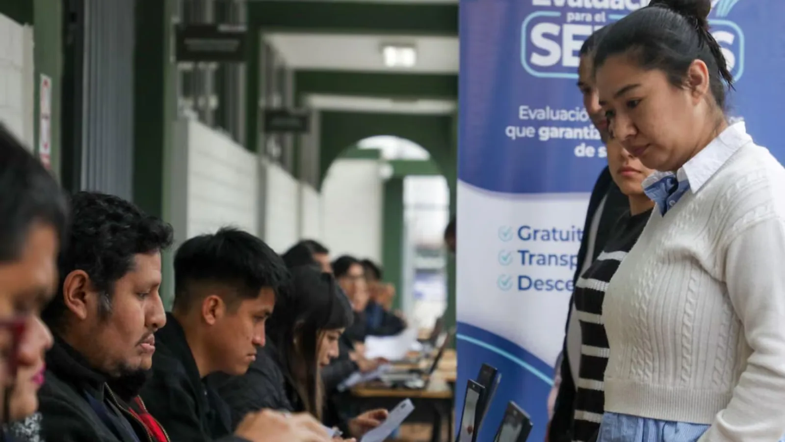 Candidatos al SERUMS 2026-I en jornada oficial de evaluación MINSA · mesa de acreditación con personal revisando documentos, banner institucional 'Evaluación para el SERUMS · gratuito, transparente, descentralizado' al fondo